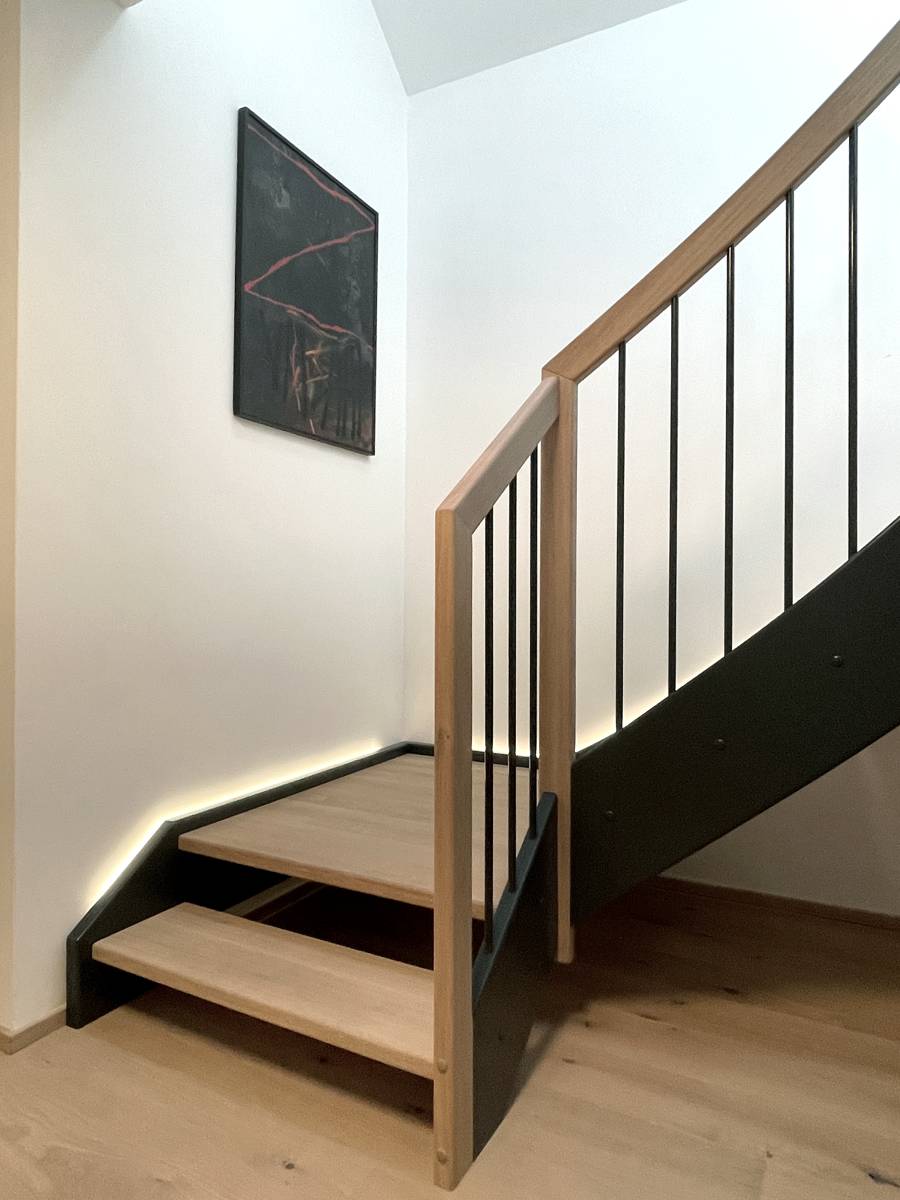 Treppe aus hellen Holzelementen, schwarzem Stahlgeländer und hölzernem Handlauf. an der Wand entlang verläuft eine moderne, indirekte Beleuchtung.