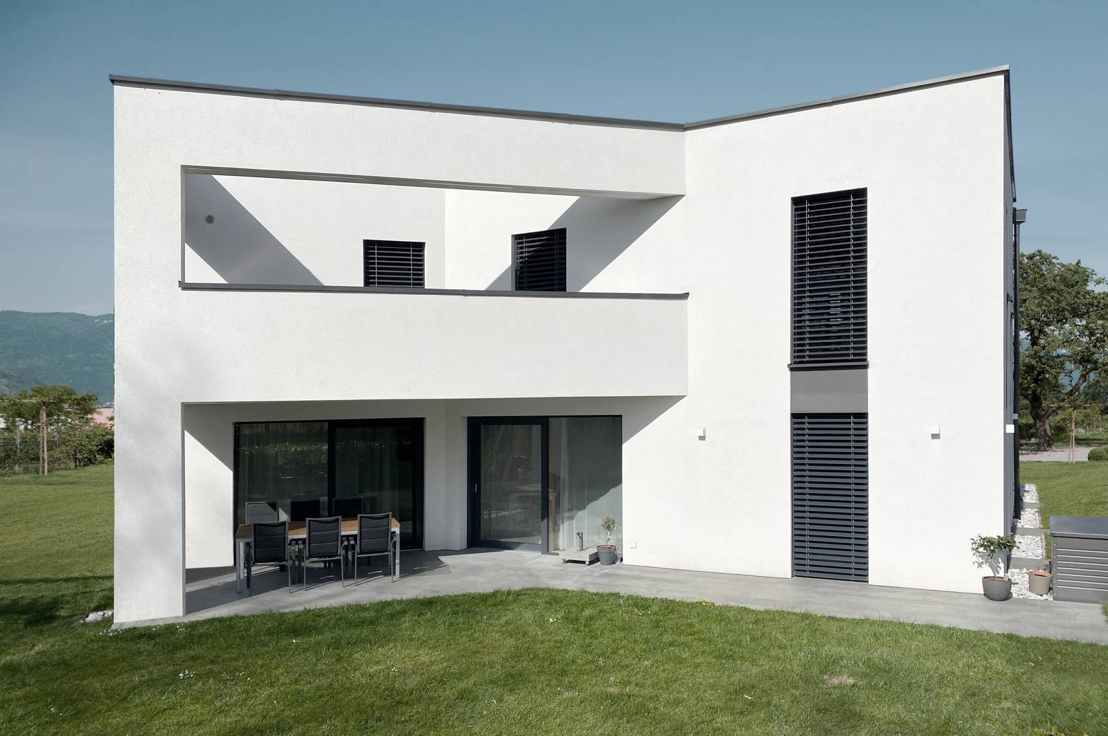 Außenansicht eines modernen, weißen Hauses mit anthrazitfarbenen Fensterelementen und Jalousien an sonnigen Tag.